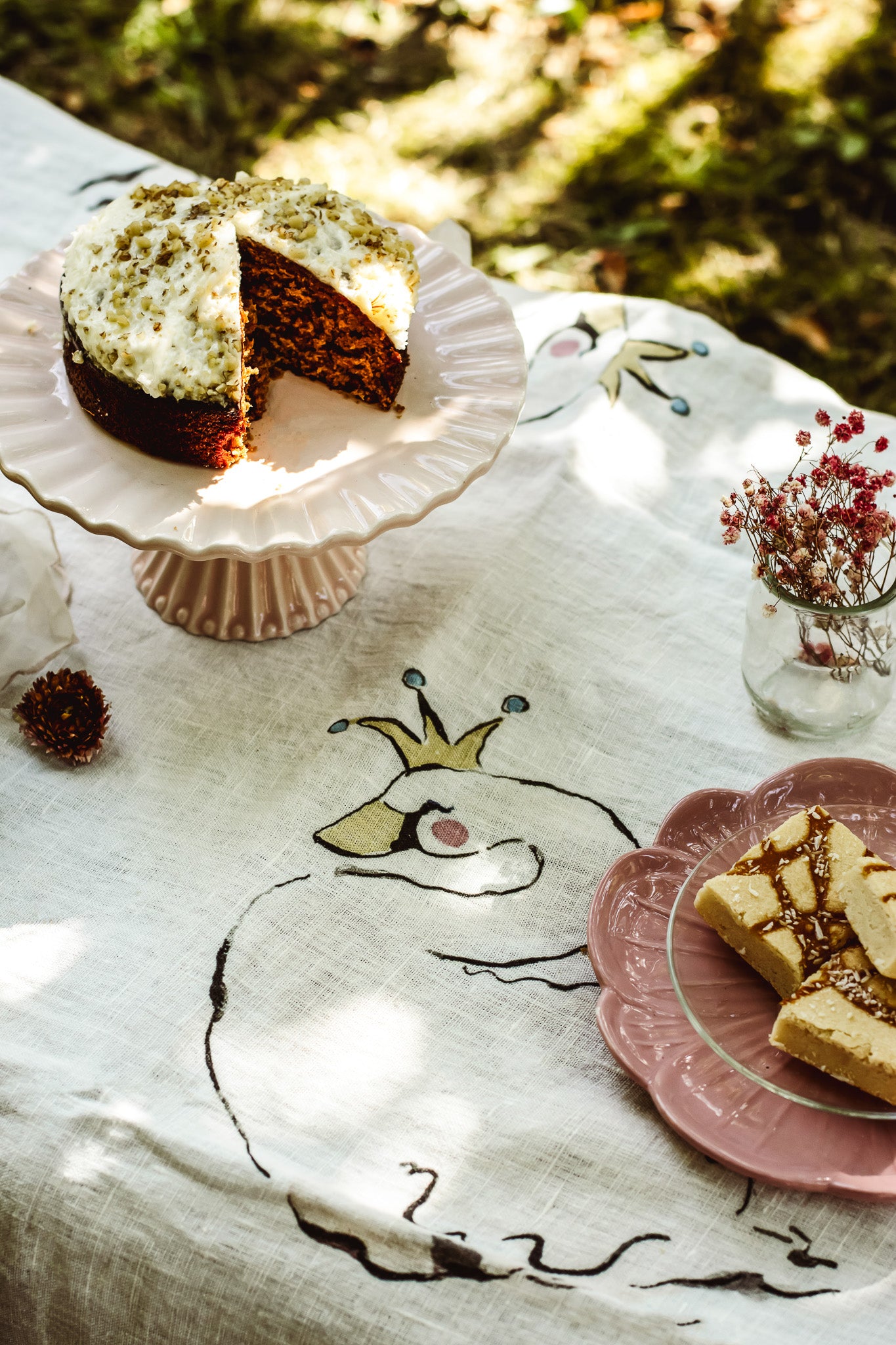 A styled outdoor table scene with a frosted cake on a pedestal plate, sliced pieces on a pink plate, and small dried flowers, all arranged on a white embroidered tablecloth with a whimsical animal design.