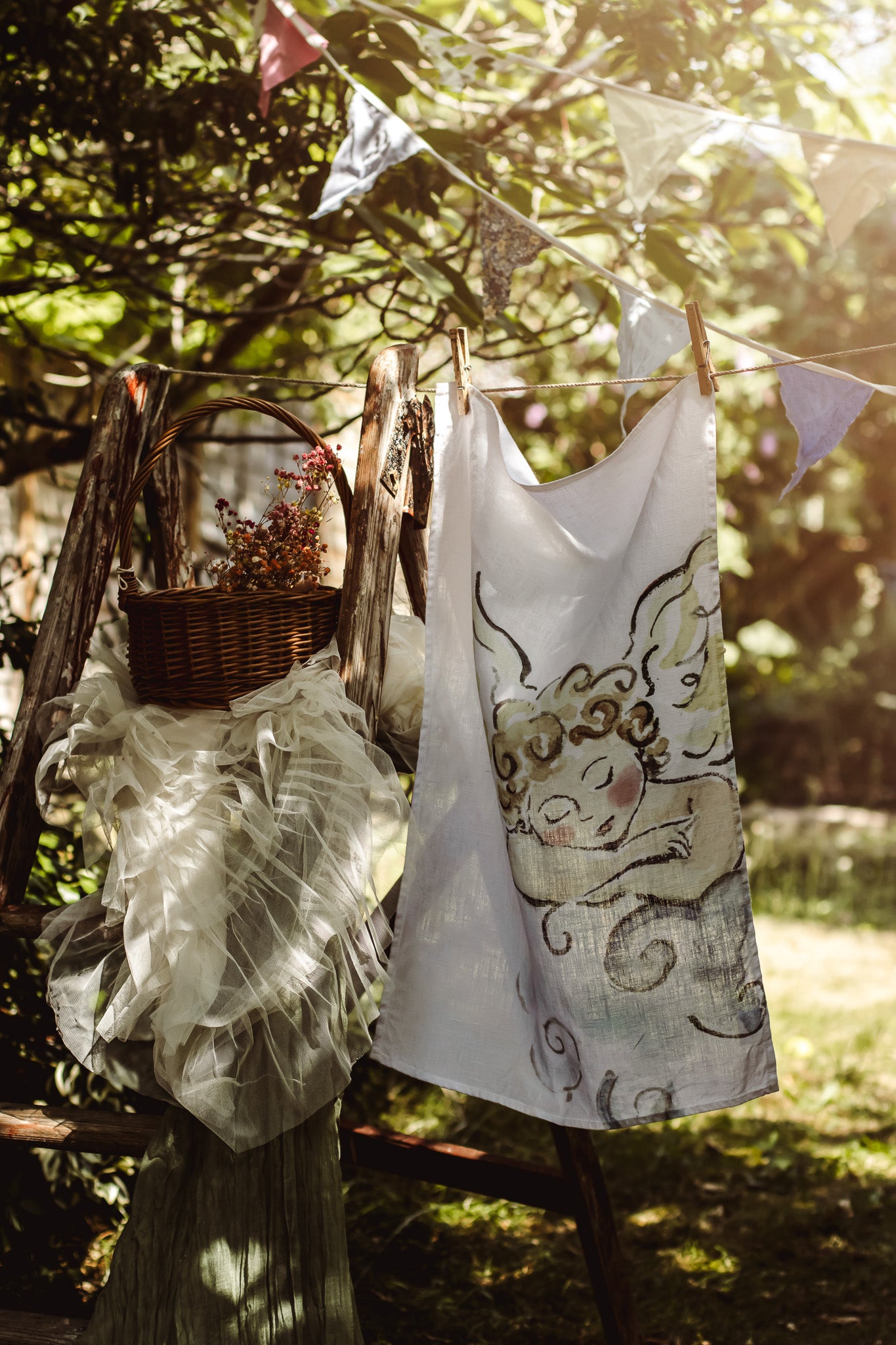 A white sleeveless top with a whimsical cartoon character print hangs on a clothesline outdoors, alongside a sheer white ruffled dress or skirt and a wicker basket with dried flowers, set against a sunlit forest background.