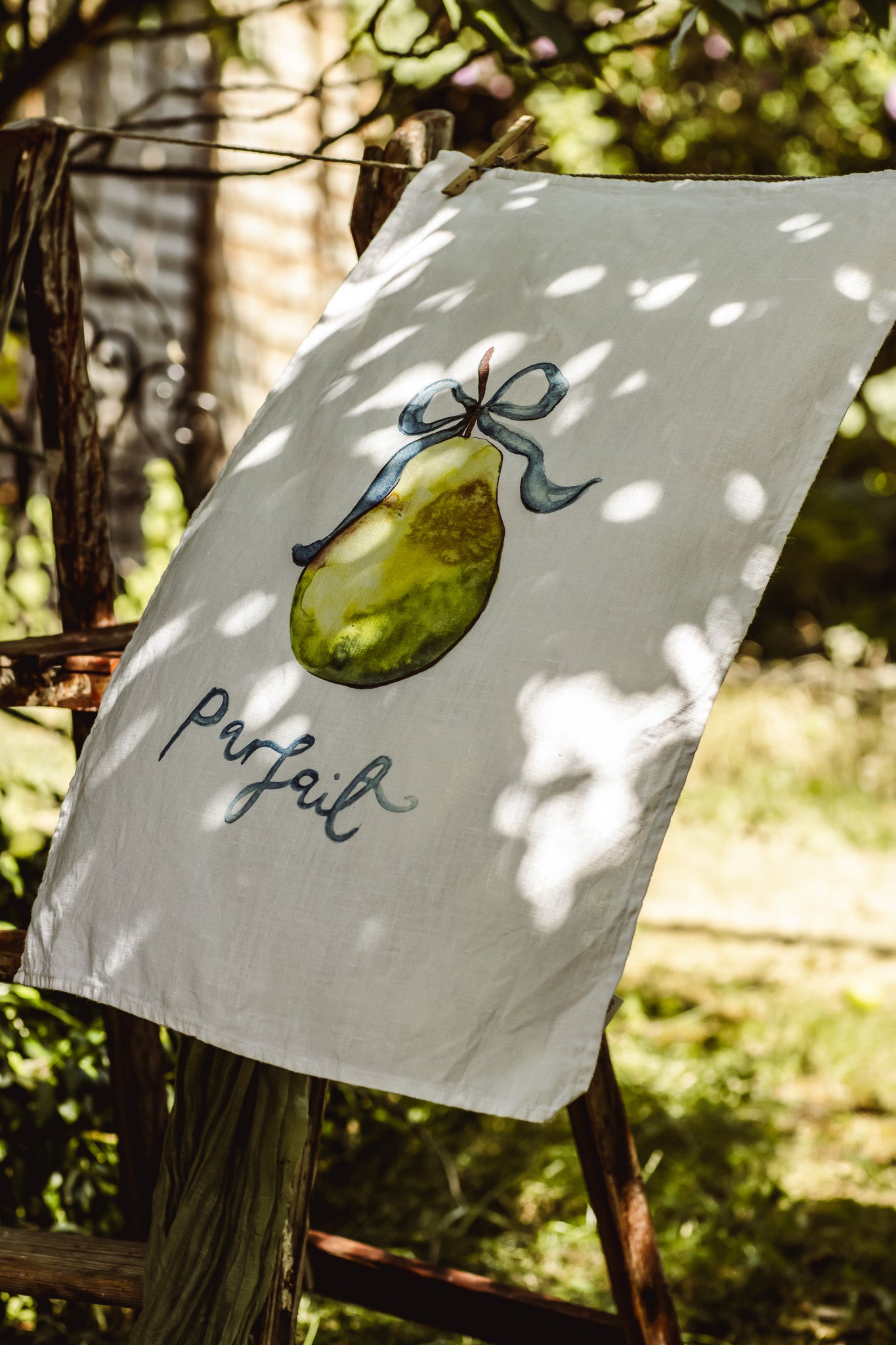 A white fabric tea towel or garden flag hanging outdoors on a rustic wooden fence. The white fabric features a painted green pear tied with a blue ribbon and the word 'parfait' in black cursive script.