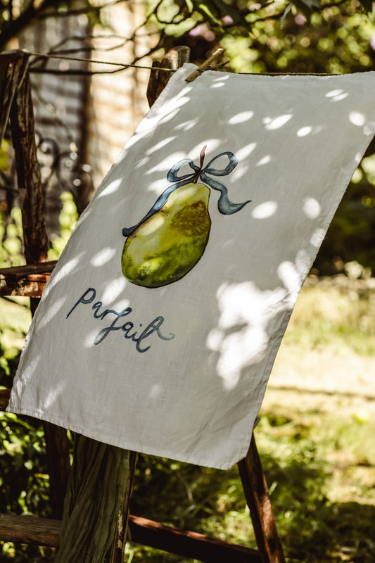 A white fabric tea towel or garden flag hanging outdoors on a rustic wooden fence. The white fabric features a painted green pear tied with a blue ribbon and the word 'parfait' in black cursive script.