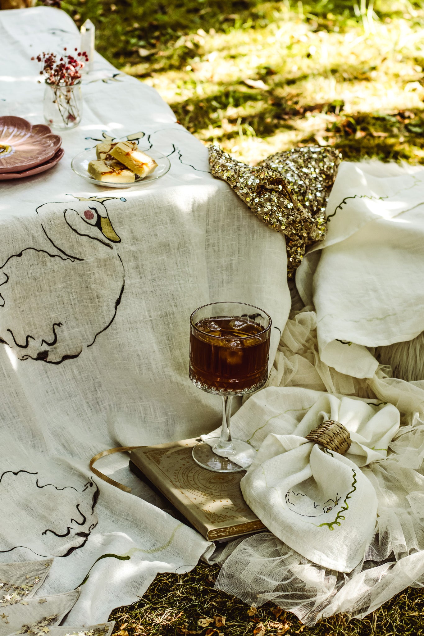 A styled outdoor picnic scene with a white embroidered linen tablecloth featuring a goose design, draped over a table with plates, a glass of iced dark beverage, a gold sequined bag, and a book on grass.